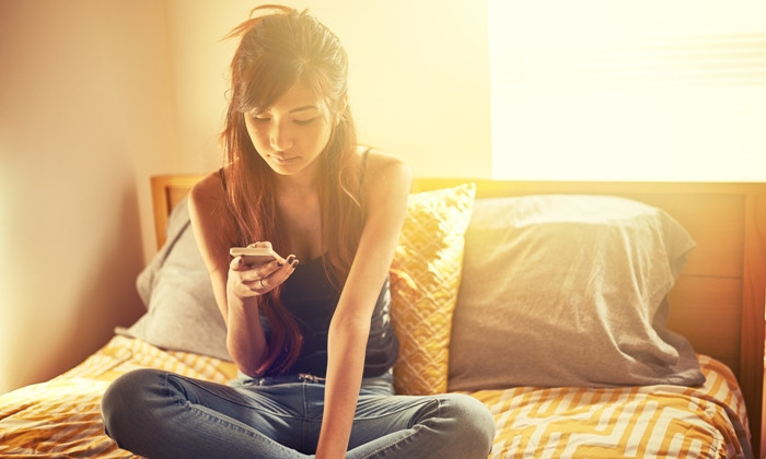 tech savvy asian teen girl using smart phone while sitting on bed in room. Image has lens flare and warm toning.